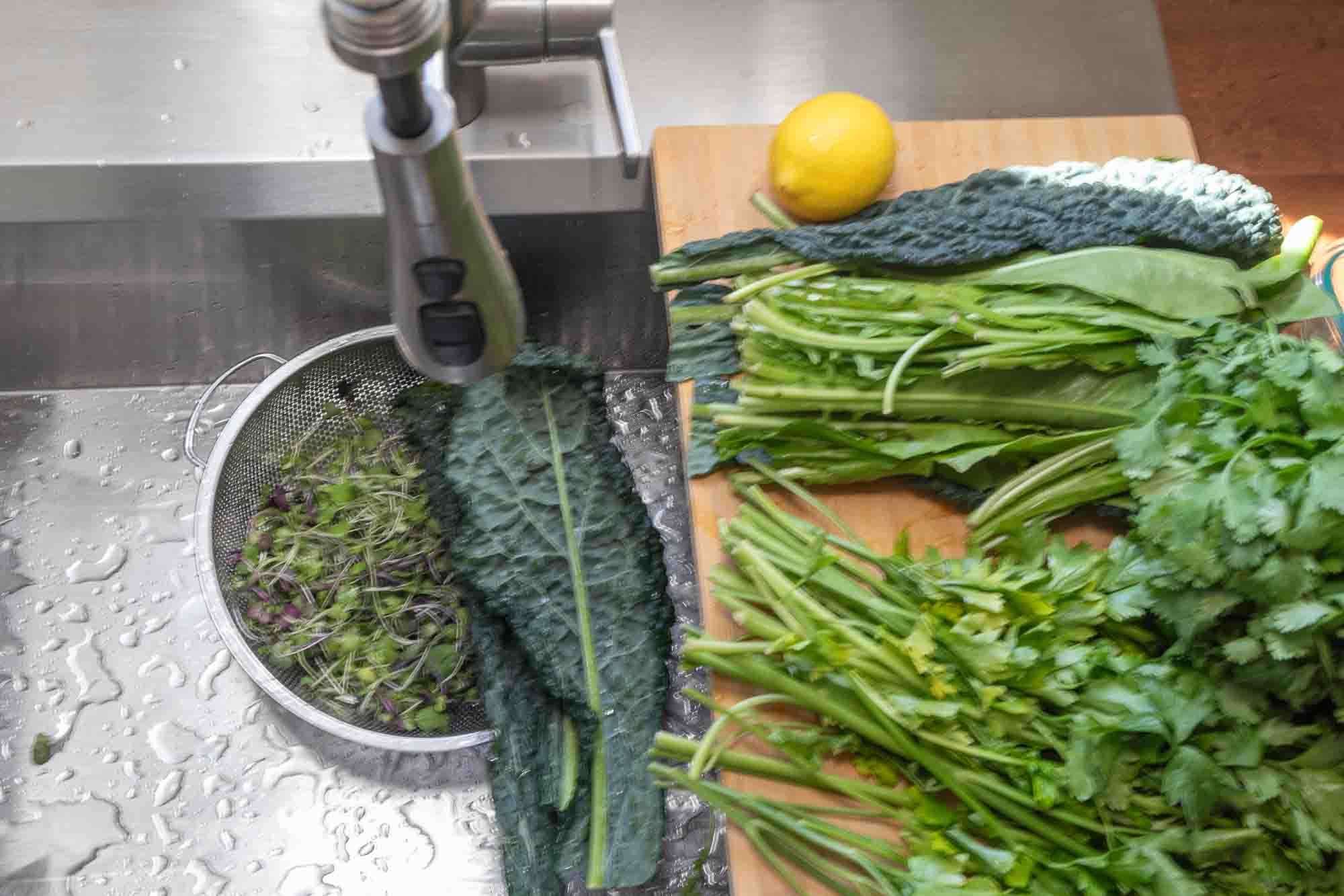 Process shot of greens in a colander being washed with more greens laid out on a cutting board in the background.